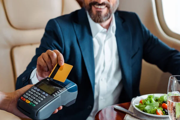 Enfoque selectivo de sonriente hombre de negocios pagando con tarjeta de crédito cerca de ensalada y copa de champán en avión - foto de stock