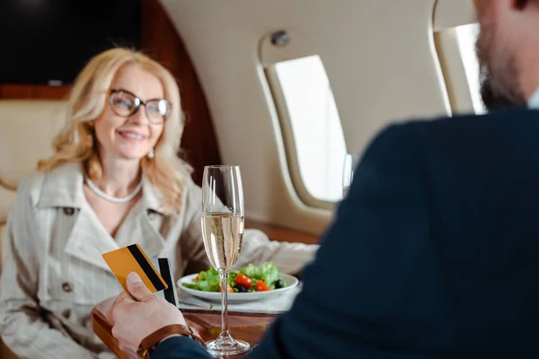 Selective focus of businessman holding credit cards near smiling businesswoman, glasses of champagne and salad in airplane — Stock Photo