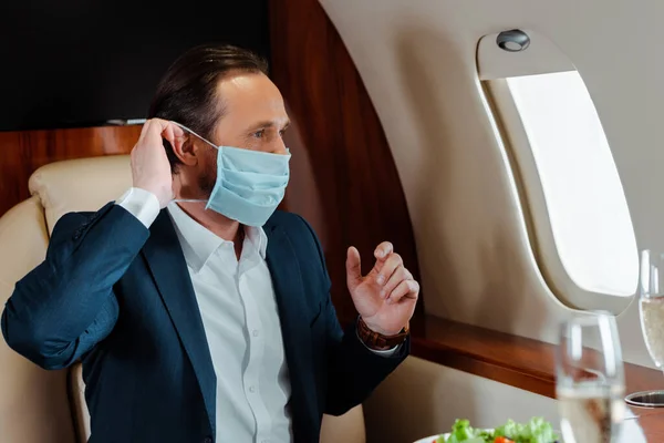 Focus selettivo dell'uomo d'affari che indossa una maschera medica vicino a champagne e insalata sul tavolo in aereo — Foto stock