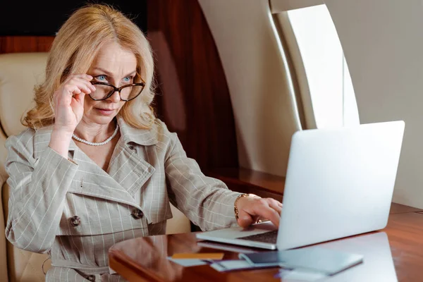 Selective focus of businesswoman holding eyeglasses while using laptop near credit cards and passports with air tickets on table in airplane — Stock Photo