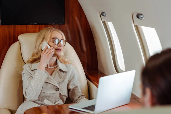 Focus selettivo della donna d'affari che parla su smartphone vicino laptop e uomo d'affari in aereo — Foto stock