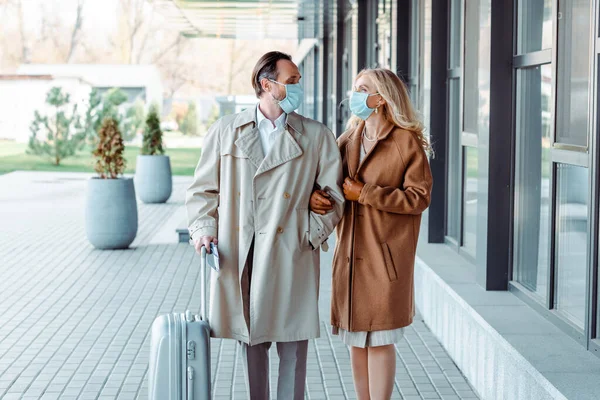 Couple d'affaires en masques médicaux se regardant tout en marchant avec une valise dans la rue urbaine — Photo de stock