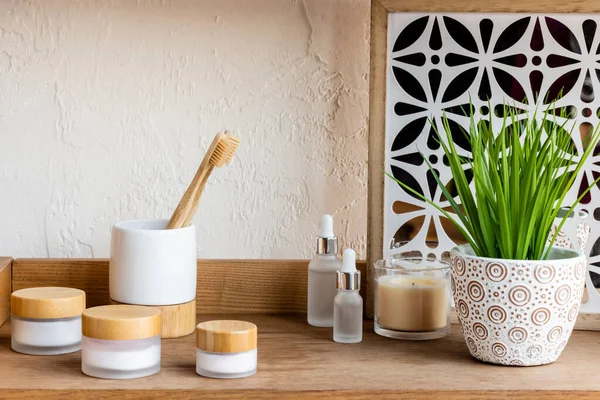 Estante de madera con cepillos de dientes, contenedores y botellas cerca de la vela y la planta verde - foto de stock