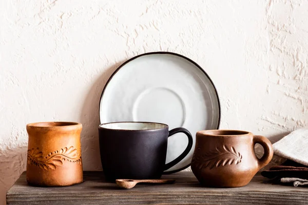 Cuchara de madera cerca de tazas de arcilla y plato en la cocina - foto de stock