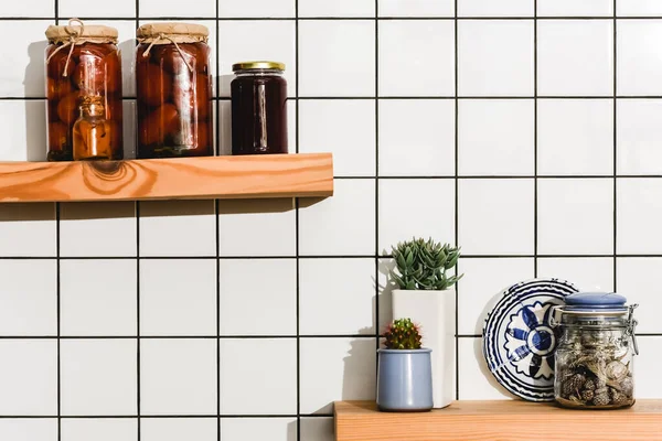 Canned tomatoes in jars near green plants and plate with ornate on shelves — Stock Photo