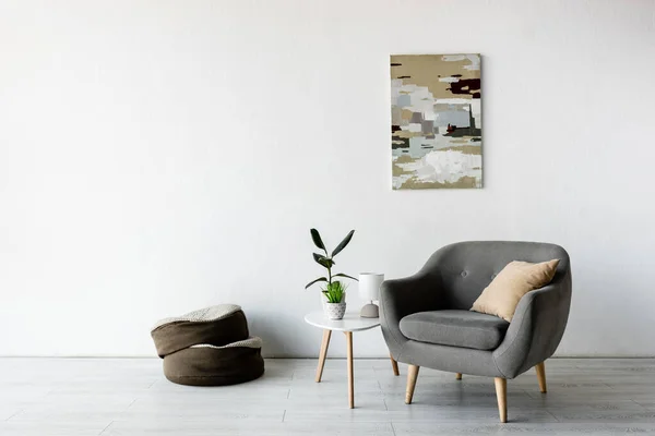 Cómodo sillón y almohada cerca de la mesa de centro con plantas verdes, lámpara y pintura en la pared en la sala de estar - foto de stock