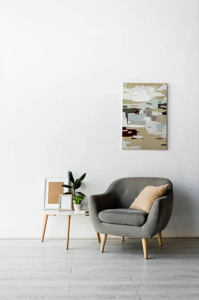 Sillón gris cerca de la mesa de centro con marcos y plantas verdes cerca de la pintura en la pared en la sala de estar moderna - foto de stock