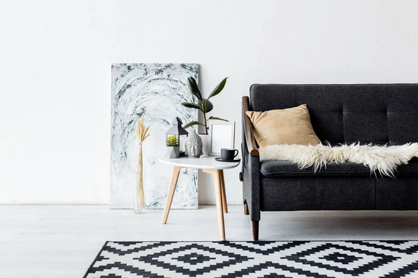 Sofá moderno perto de plantas e mesa de café com lâmpada vintage, estatueta de cabeça e xícara na sala de estar com tapete — Fotografia de Stock