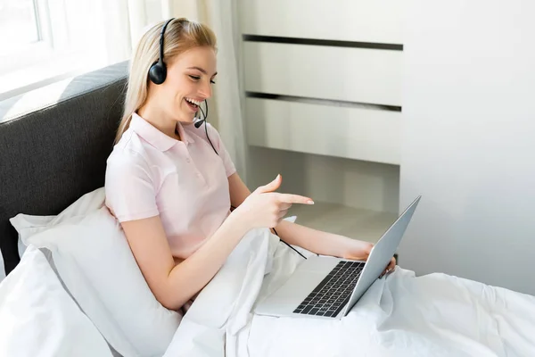 Alegre freelancer en auriculares apuntando con el dedo al portátil en el dormitorio - foto de stock