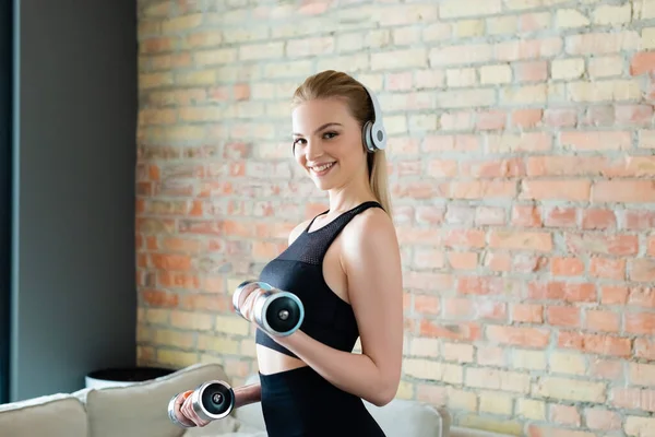 Chica feliz y deportiva en auriculares inalámbricos haciendo ejercicio con mancuernas en casa - foto de stock