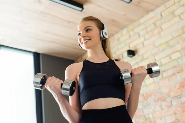 Vista basso angolo di ragazza allegra e sportiva in cuffie wireless esercizio con manubri a casa — Foto stock