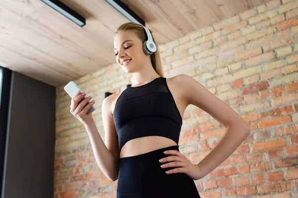 Low angle view of happy sportswoman in wireless headphones using smartphone and standing with hand in pocket — Stock Photo