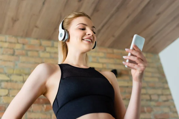 Low angle view of cheerful sportswoman in wireless headphones using smartphone and standing with hand in pocket — Stock Photo