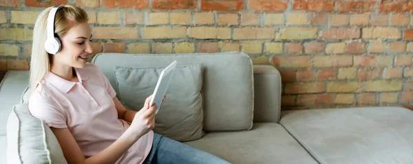 Plano panorámico de la mujer sonriente en auriculares inalámbricos utilizando tableta digital en la sala de estar - foto de stock