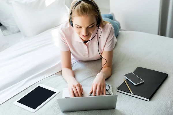 Happy freelancer in headset using laptop near gadgets with blank screen on bed — Stock Photo