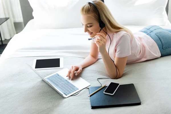 Alegre freelancer en auriculares usando portátil cerca de aparatos con pantalla en blanco en la cama - foto de stock