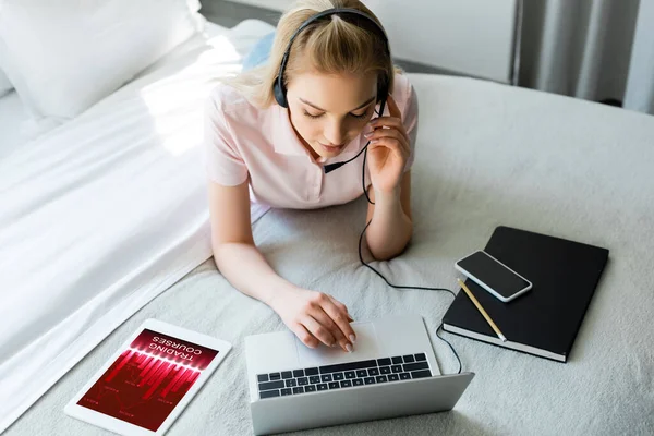 Jeune pigiste dans casque à l'aide d'un ordinateur portable près de tablette numérique avec des cours de trading et smartphone avec écran blanc sur le lit — Photo de stock