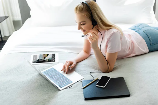 Cheerful girl in headset using laptop near digital tablet with booking app and smartphone with blank screen on bed — Stock Photo