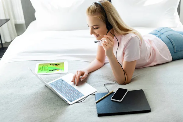 Cheerful freelancer in headset using laptop near digital tablet with online shopping app and smartphone with blank screen on bed — Stock Photo