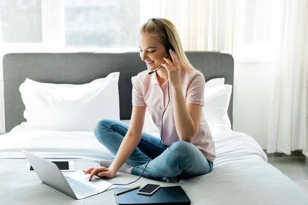 Operador alegre no fone de ouvido usando laptop perto de gadgets com tela em branco na cama — Fotografia de Stock