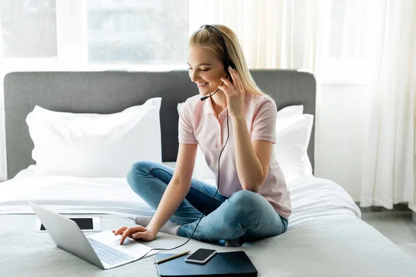 Operador alegre en auriculares con portátil cerca de aparatos con pantalla en blanco en la cama - foto de stock