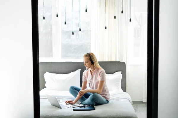 Beautiful operator in headset using laptop near gadgets with blank screen on bed — Stock Photo