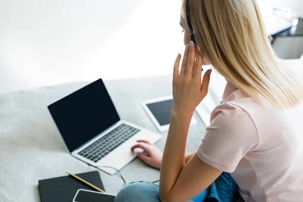 Foyer sélectif de freelance toucher casque près d'un ordinateur portable et tablette numérique avec écran blanc sur le lit — Photo de stock