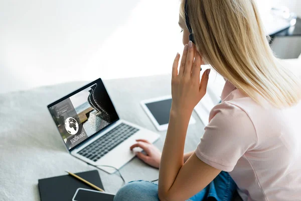 Enfoque selectivo de freelancer tocando auriculares cerca de la computadora portátil con sitio web de reserva y gadgets con pantalla en blanco en la cama - foto de stock