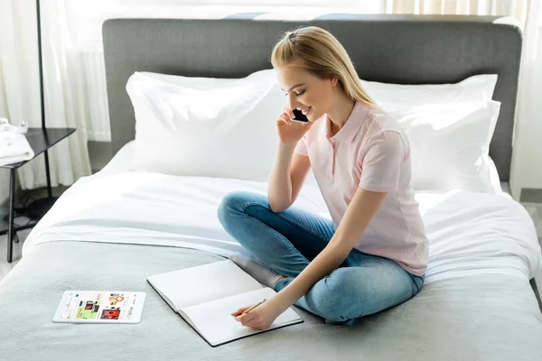 KIEW, UKRAINE - 8. April 2020: Glückliche Frau blickt auf Notebook und spricht auf Smartphone neben digitalem Tablet mit ebay-App im Schlafzimmer — Stockfoto
