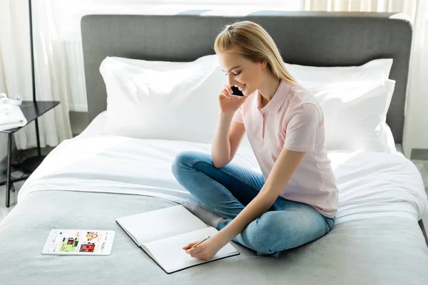 KYIV, UKRAINE - 8 AVRIL 2020 : femme heureuse regardant un ordinateur portable et parlant sur un smartphone près d'une tablette numérique avec une application ebay dans la chambre — Photo de stock