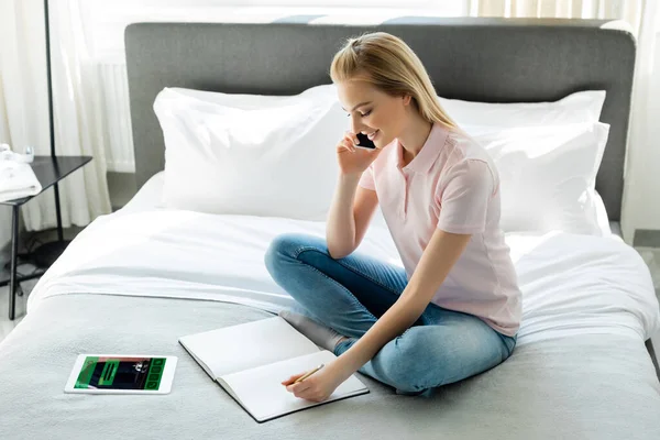 Mujer feliz mirando el cuaderno en blanco y hablando en el teléfono inteligente cerca de la tableta digital con aplicación de reserva en la pantalla en el dormitorio - foto de stock