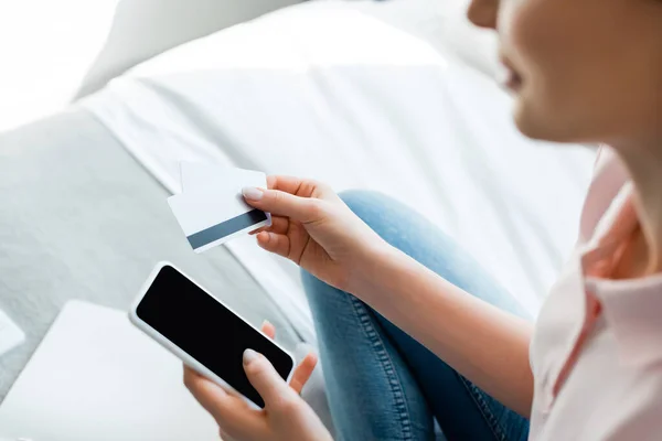 Enfoque selectivo de la mujer que sostiene el teléfono inteligente con pantalla en blanco y tarjetas de crédito en el dormitorio - foto de stock