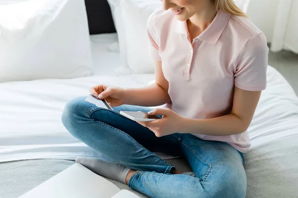Cropped view of happy woman holding smartphone and credit cards in bedroom — Stock Photo