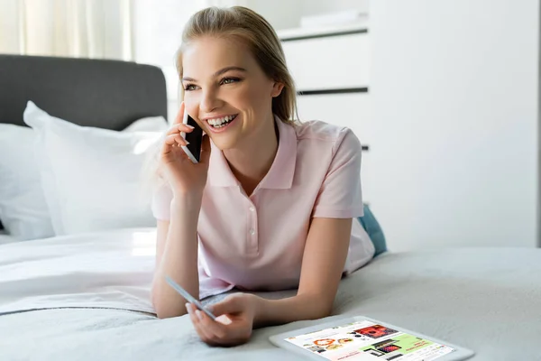 KYIV, UCRANIA - 8 DE ABRIL DE 2020: mujer feliz hablando en el teléfono inteligente y la celebración de la tarjeta de crédito cerca de la tableta digital con la aplicación ebay en el dormitorio - foto de stock