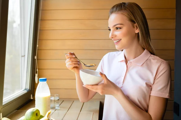 Femme heureuse tenant cuillère avec des flocons de maïs près de la bouteille avec du lait et des fruits — Photo de stock