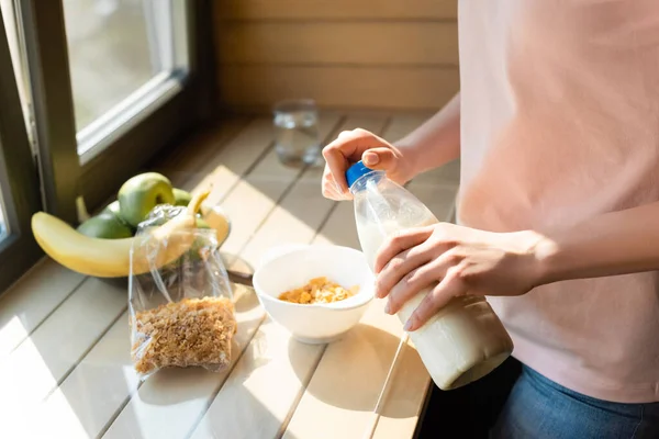 Cropped view of woman holding bottle with milk near cornflakes in bowl and fruits — Stock Photo