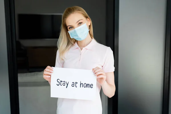 Junge Frau mit medizinischer Maske hält Papier mit dem Schriftzug Stay at Home in der Hand — Stockfoto