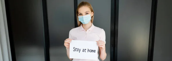 Tiro panorâmico de mulher jovem em máscara médica segurando papel com estadia em casa lettering — Fotografia de Stock