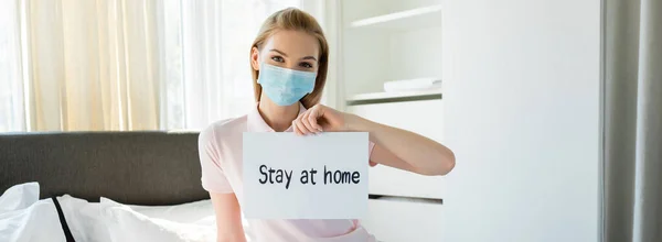 Tiro panorâmico de jovem mulher em máscara médica segurando papel com estadia em casa lettering no quarto — Fotografia de Stock