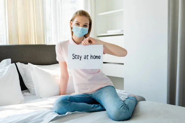 Mujer joven con máscara médica sosteniendo papel con letras de estancia en casa mientras está sentada en la cama - foto de stock