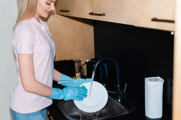 Felice donna in guanti di gomma piatto di lavaggio vicino al rotolo di tessuto in cucina — Foto stock