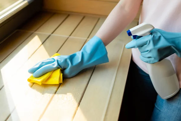 Vue recadrée de la jeune fille dans des gants en caoutchouc tenant bouteille de pulvérisation et chiffon près de la surface en bois — Photo de stock