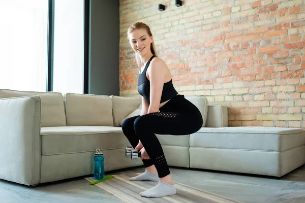 Felice sportswoman facendo esercizio squat con manubri vicino bottiglia di sport in soggiorno — Foto stock