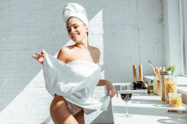 Lächelnde sexy Frau in Höschen und Handtüchern steht neben einem Glas Wein auf der Arbeitsplatte in der Küche — Stockfoto