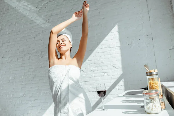 Chica alegre envuelta en toallas de pie cerca de un vaso de vino en la encimera en la cocina - foto de stock