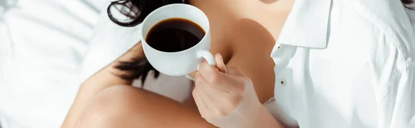 Cropped view of sexy woman in shirt and bra holding coffee on bed, panoramic shot — Stock Photo
