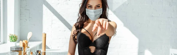 Tiro panorâmico de mulher sexy no sutiã e máscara médica apontando com o dedo para a câmera na cozinha — Fotografia de Stock