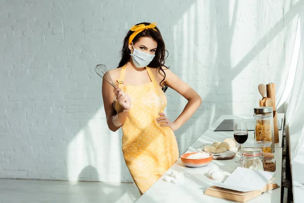 Menina em máscara médica e avental segurando batedor perto da massa e ingredientes na cozinha — Fotografia de Stock
