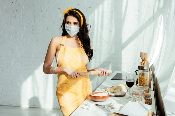 Nacktes Mädchen in Schürze und medizinischer Maske mit Nudelholz beim Kochen in der Küche — Stockfoto