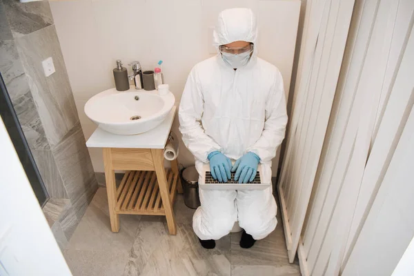 Hombre en traje de materiales peligrosos, máscara médica, guantes de látex y gafas usando el ordenador portátil en el baño - foto de stock